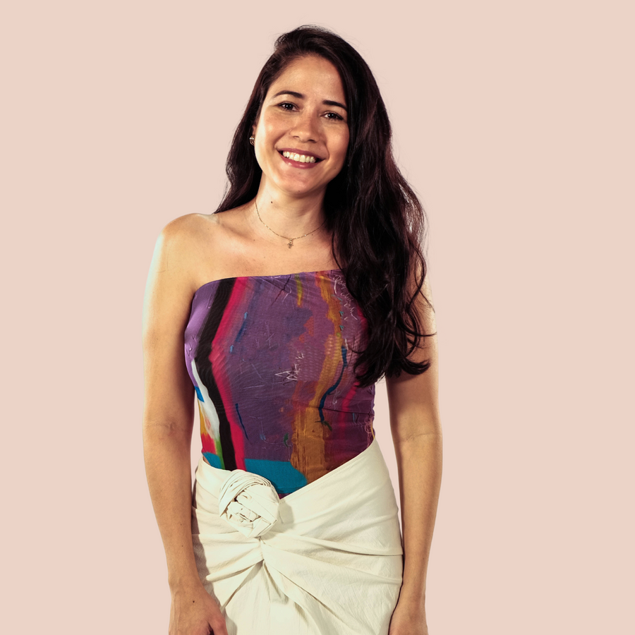 A Hispanic woman smiling. The woman has long dark hair and tan skin color. She is wearing a multicolor top and a beige color skirt. The text next to the image states "Proudly woman owned learn about our co-founder's journey to discover matcha". Greenboxed is co founded and led by a latin / latinx woman minority own and it is a wellness brand owned by a woman. The brand is now located in Pittsburgh, PA. Proudly providing organic unsweetened matcha.